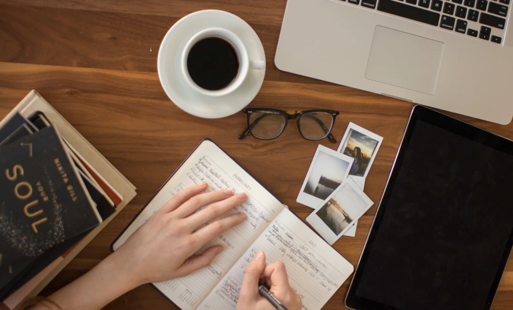 Person writing in a note book at a desk with a cup of coffee, a pen, some books, and a laptop