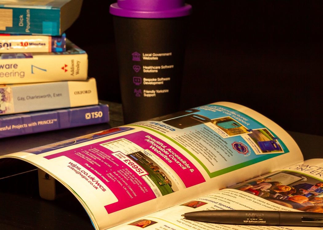The Clerk Magazine open displaying the WJPS advert, in front of a WJPS travel mug and some software development books